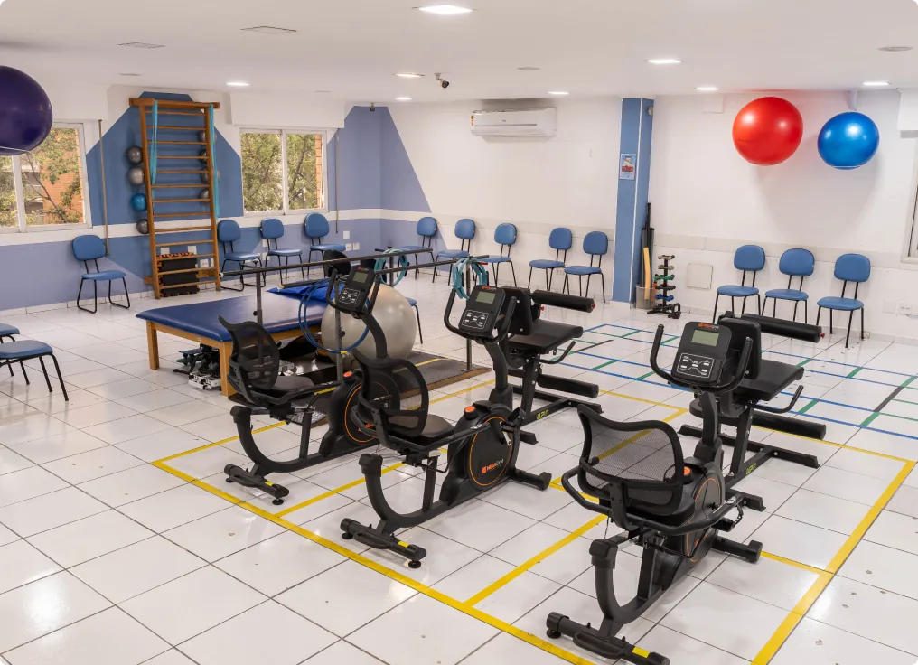 Vista da sala de fisioterapia com aparelhos de exercícios e marcações coloridas no piso.